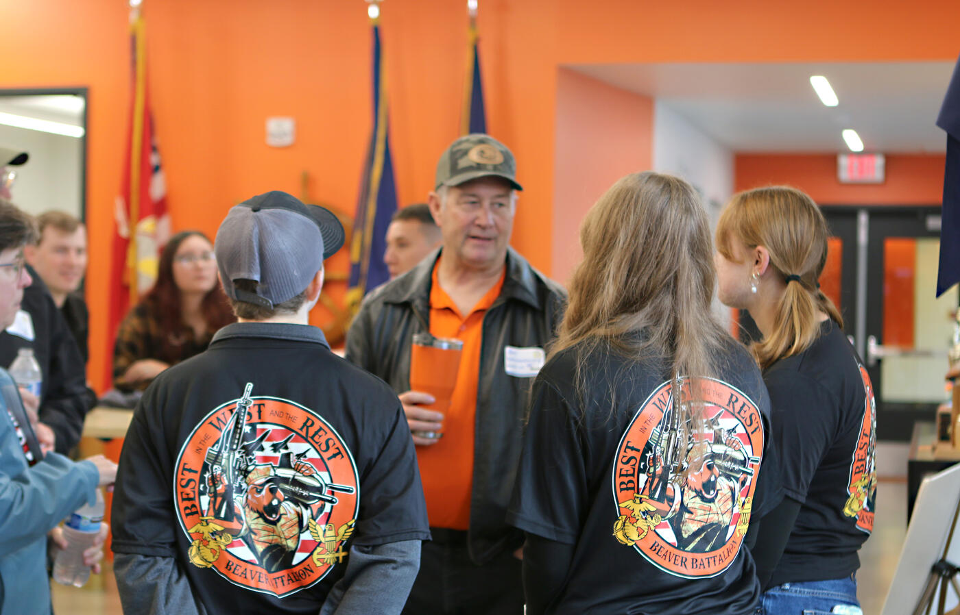 Older gentleman talking to a round of students, in an orange room. 