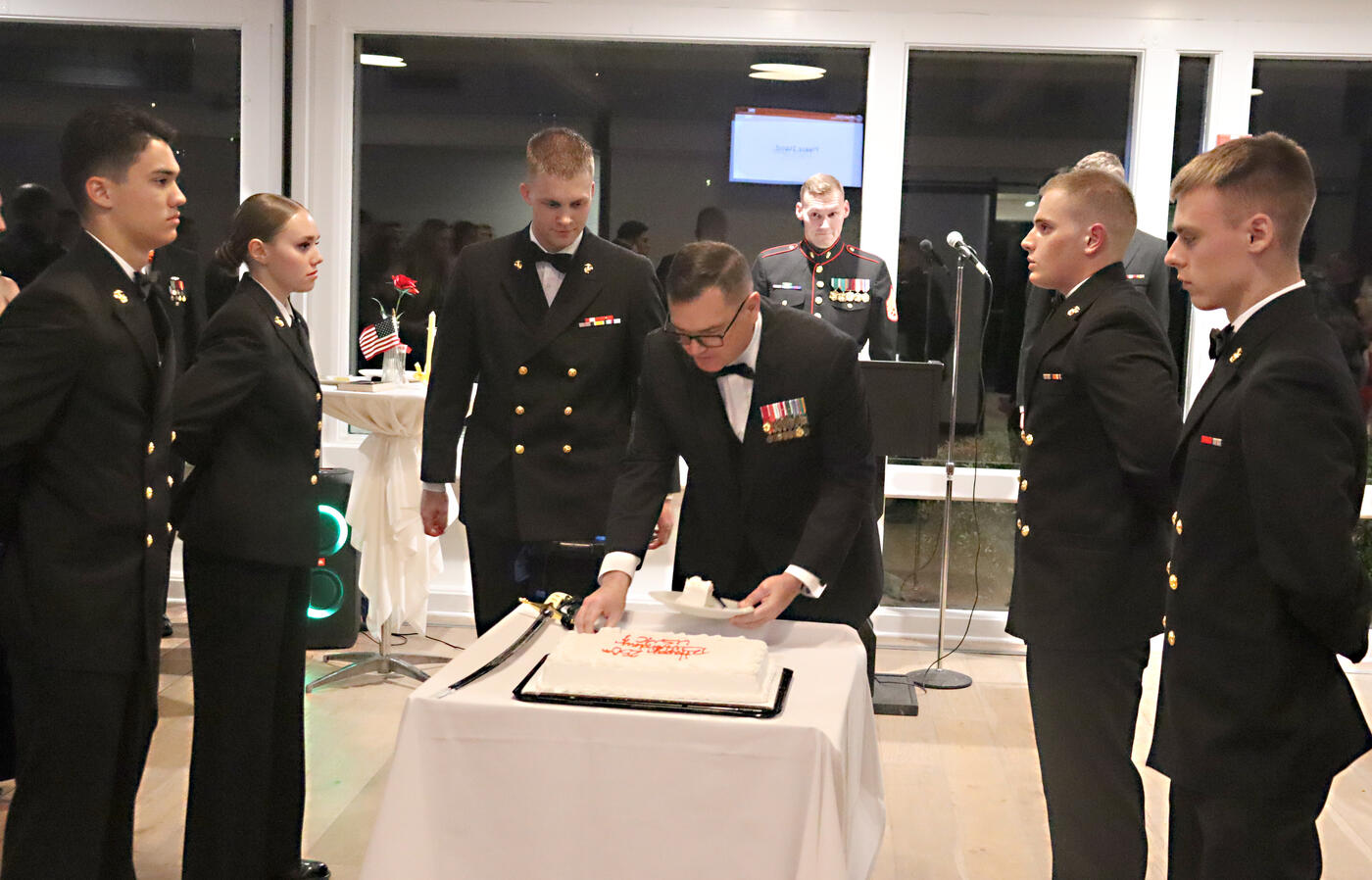 Student in uniform standing around as a older man cuts a piece of cake. 